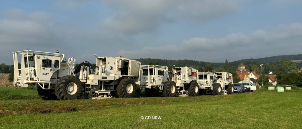 Kolonne von Vibro-Trucks auf grüner Wiese mit leicht bewölktem Himmel.