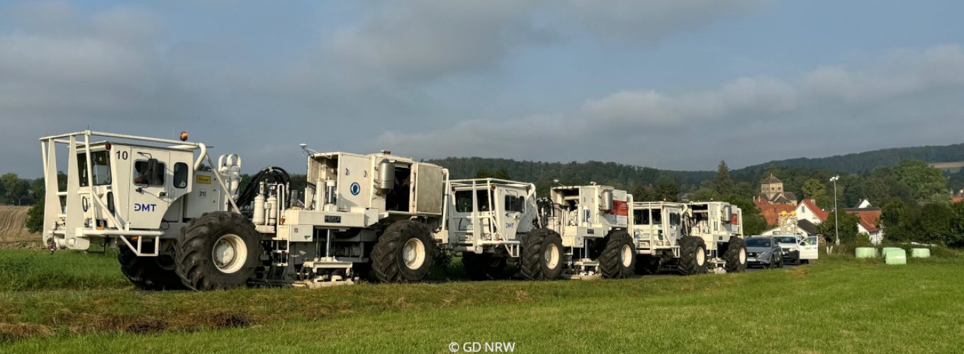Kolonne von Vibro-Trucks auf grüner Wiese mit leicht bewölktem Himmel.